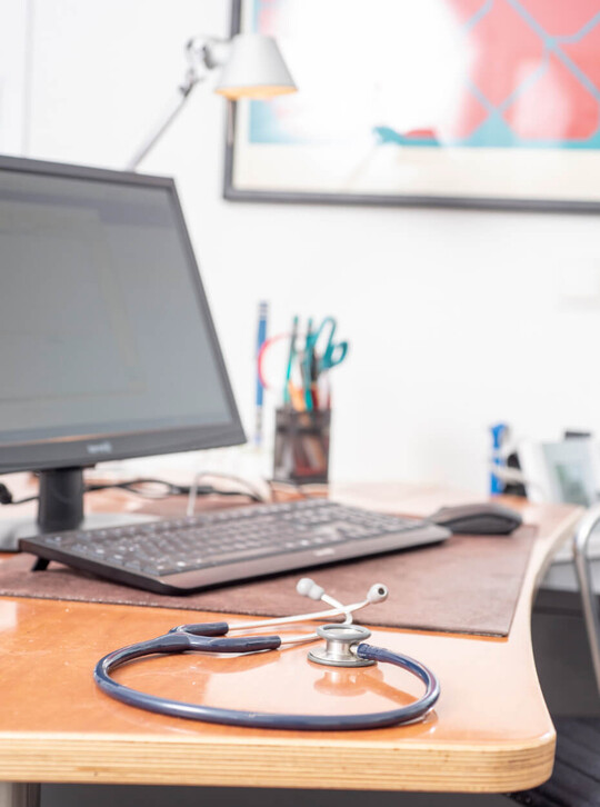 Schreibtisch mit Computer, Stift und medizinischen Unterlagen in einer Arztpraxis